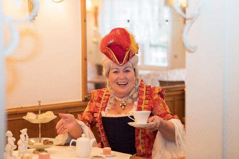 Frau in historischer Kleidung mit rotem Hut und Perlenkette genie&szlig;t Kaffee in einem Caf&eacute;. Auf dem Tisch stehen Geb&auml;ck und eine Kaffeekanne., &copy; Fotografin: Jasmin Schuler