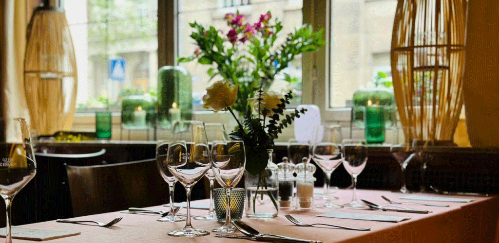 Ein stilvoll gedeckter Tisch in einem Café mit Weingläsern, Besteck und Blumenarrangement vor einem Fenster., © FreCla Gastronomiebetriebe GmbH Ein stilvoll gedeckter Tisch in einem Café mit Weingläsern, Besteck und Blumenarrangement vor einem Fenster., © FreCla Gastronomiebetriebe GmbH