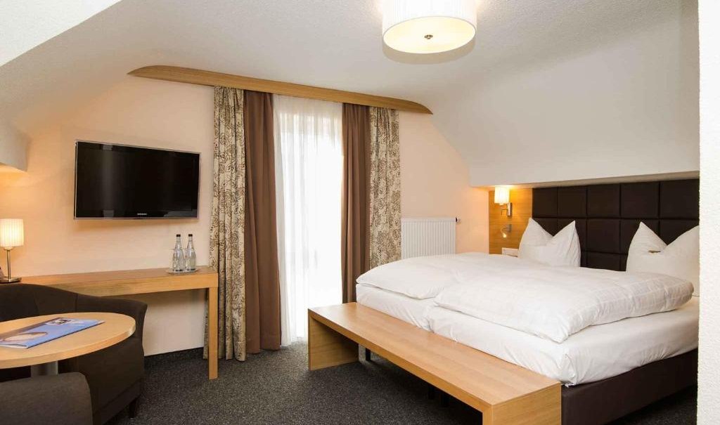 Modernes Doppelzimmer mit großem Bett, Flachbildfernseher, Sitzbereich und Tisch. Helle Wände, Vorhänge und Teppichboden., © Nödinger Gastro GmbH