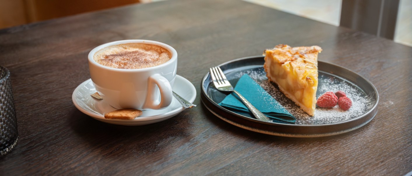 Ein Cappuccino mit Keks und ein St&uuml;ck Apfelkuchen mit Himbeeren auf einem Tisch. Perfekt f&uuml;r eine gem&uuml;tliche Kaffeepause., &copy; Lunar