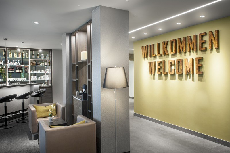 Moderne Lobby mit Bar, Sitzbereich und großem "Willkommen"-Schild an der Wand. Elegante Einrichtung mit Barhockern und Stehlampe., © TOMAS