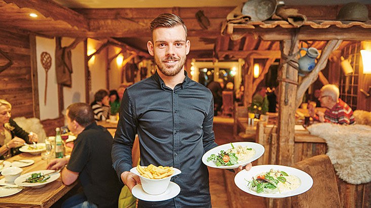 Ein Kellner im Restaurant Himmelreich Schorndorf serviert zwei Teller mit Pasta und eine Schüssel Pommes. Im Hintergrund sitzen Gäste an Holztischen., © Restaurant Himmelreich Schorndorf