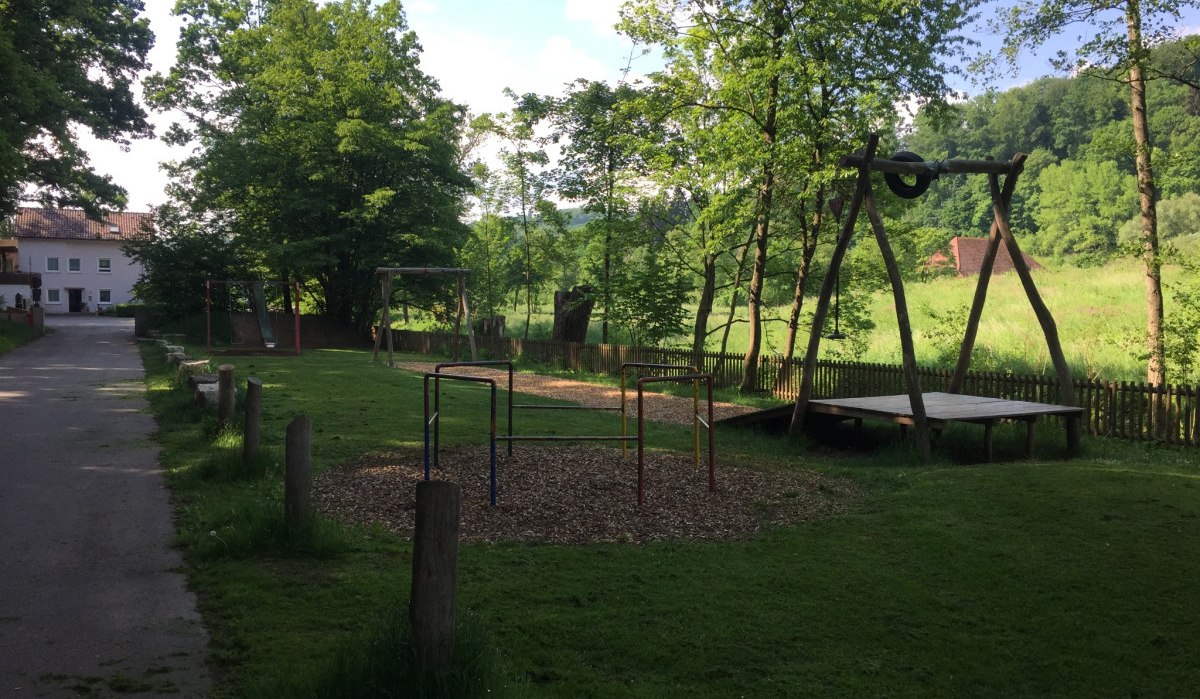 Ein ruhiger Spielplatz mit Schaukeln und Klettergerüst, umgeben von Bäumen und Wiesen. Ein Weg führt am Spielplatz vorbei., © www.pro-cycl.de Ein ruhiger Spielplatz mit Schaukeln und Klettergerüst, umgeben von Bäumen und Wiesen. Ein Weg führt am Spielplatz vorbei., © www.pro-cycl.de