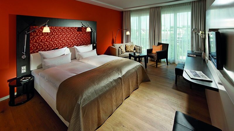 Modernes Hotelzimmer mit gro&szlig;em Bett, oranger Wand, Sofa, Schreibtisch und Fernseher. Helle Vorh&auml;nge und Holzboden schaffen eine gem&uuml;tliche Atmosph&auml;re., &copy; Parkhotel Stuttgart Messe-Airport
