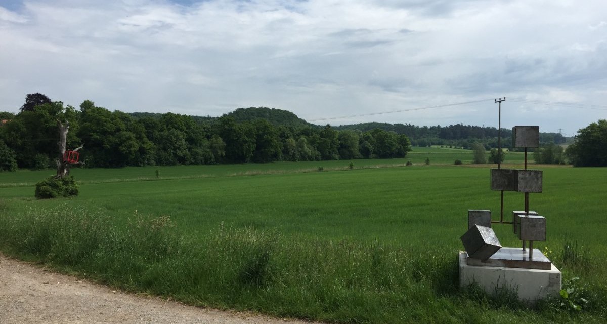 Ein Feldweg mit modernen Skulpturen in einer gr&uuml;nen Landschaft. Im Hintergrund sind B&auml;ume und H&uuml;gel zu sehen, der Himmel ist leicht bew&ouml;lkt., &copy; www.pro-cycl.de