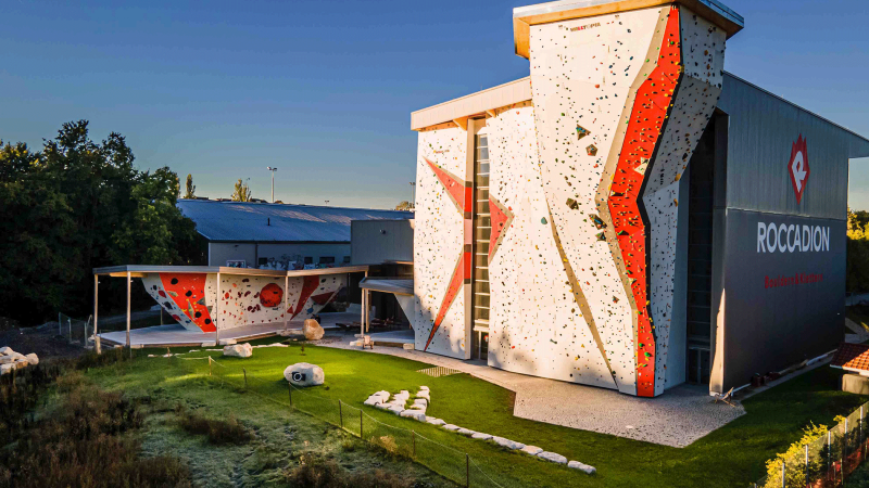 Kletterhalle mit hohen Kletterw&auml;nden und Boulderbereich im Freien, umgeben von B&auml;umen und Wiese, bei Sonnenuntergang., &copy; Roccadion, B&ouml;blingen