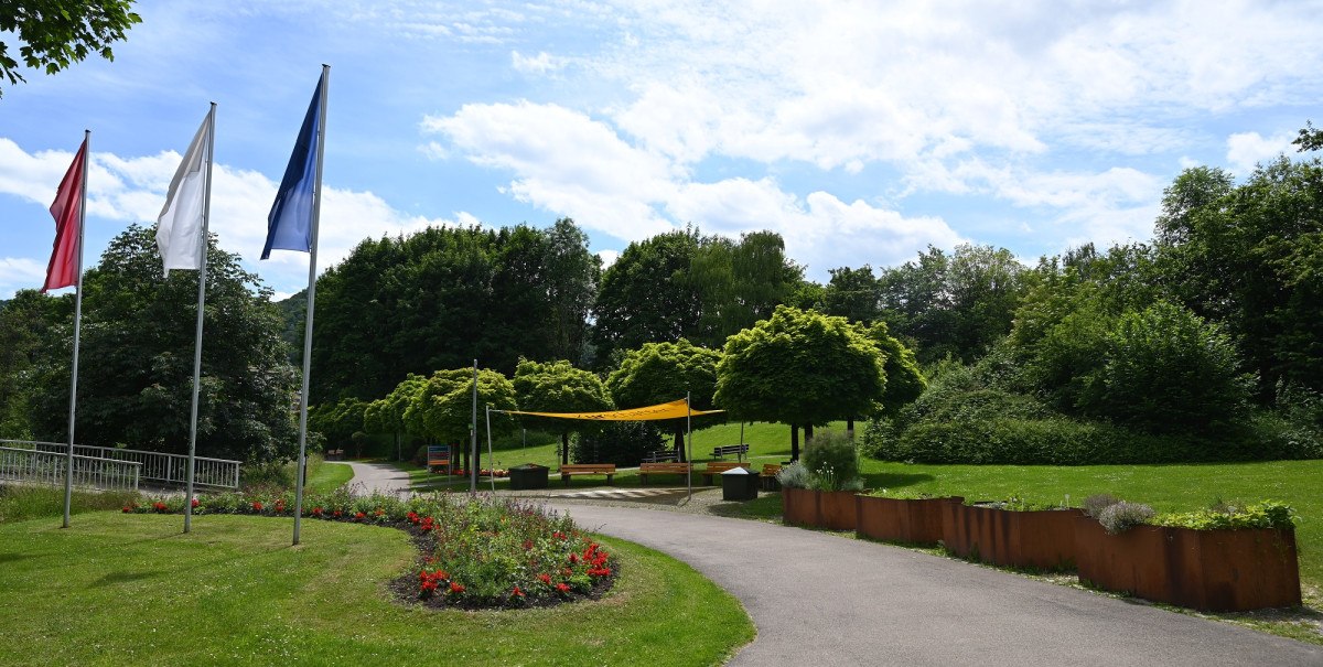 Der Kurpark Bad Urach zeigt einen Weg mit Blumenbeeten, drei Fahnen und Bäumen unter einem blauen Himmel mit Wolken.