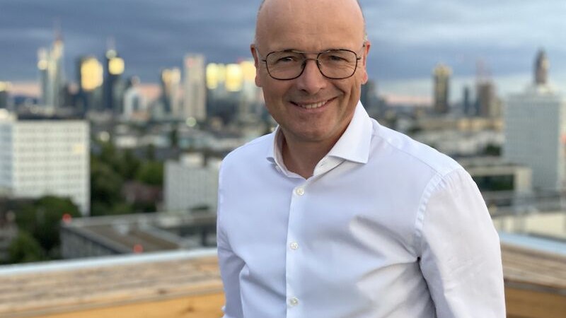 Ein Mann mit Brille und weißem Hemd steht lächelnd auf einer Dachterrasse. Im Hintergrund ist eine Stadt mit Hochhäusern zu sehen., © Stadthalle Leonberg Ein Mann mit Brille und weißem Hemd steht lächelnd auf einer Dachterrasse. Im Hintergrund ist eine Stadt mit Hochhäusern zu sehen., © Stadthalle Leonberg