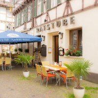 Ein Restaurant namens Ratsstube mit Außenbestuhlung unter einem blauen Sonnenschirm. Fachwerkgebäude im Hintergrund, Pflanzen schmücken den Eingangsbereich.