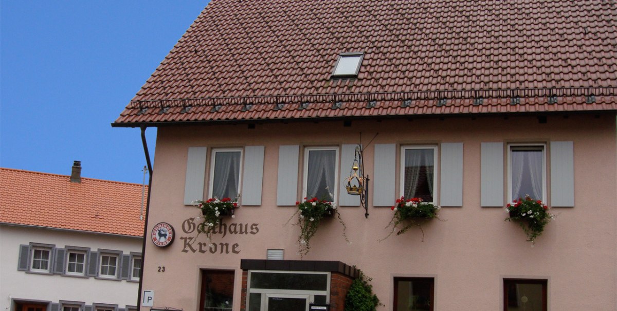 Gasthaus Krone mit blumengeschmückten Fenstern, zwei geparkte Autos davor, blauer Himmel im Hintergrund.