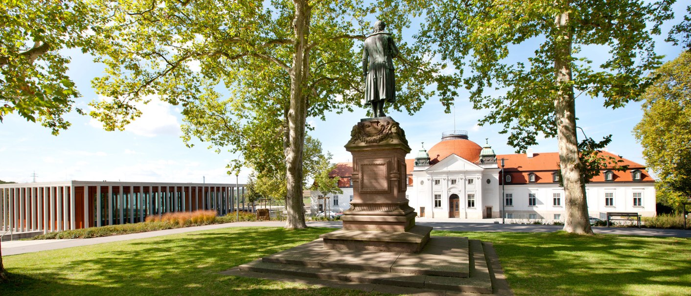 Statue auf der Schillerhöhe in Marbach am Neckar, umgeben von Bäumen. Im Hintergrund ein historisches Gebäude mit rotem Dach., © Corinna Jacobs