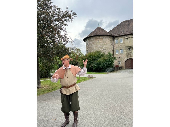 Ein Mann in historischer Kleidung gestikuliert vor einem alten Gebäude mit Rundturm. Der Himmel ist bewölkt, und Bäume umgeben den Platz., © Eigenbetrieb Tourismus und Citymanagement Schorndorf Ein Mann in historischer Kleidung gestikuliert vor einem alten Gebäude mit Rundturm. Der Himmel ist bewölkt, und Bäume umgeben den Platz., © Eigenbetrieb Tourismus und Citymanagement Schorndorf