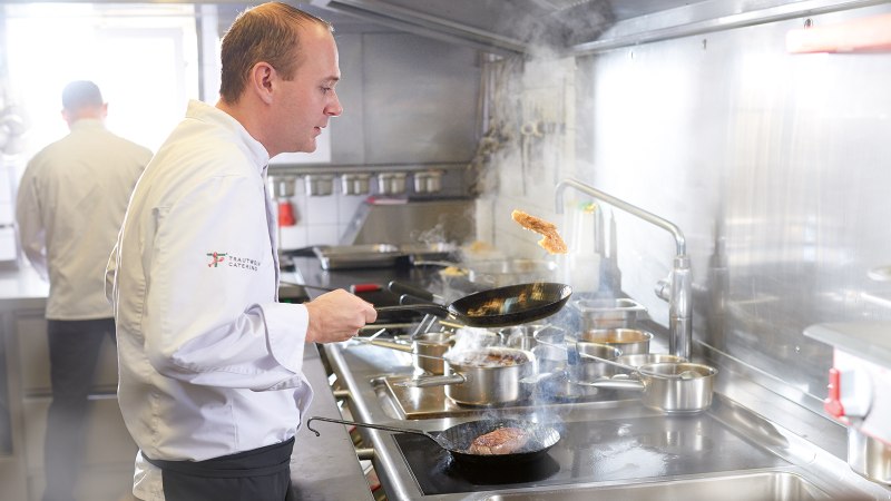 Ein Koch in weißer Uniform brät Fleisch in einer Pfanne in einer professionellen Küche. Im Hintergrund ist ein weiterer Koch zu sehen., © SMG, Jean-Claude Winkler Ein Koch in weißer Uniform brät Fleisch in einer Pfanne in einer professionellen Küche. Im Hintergrund ist ein weiterer Koch zu sehen., © SMG, Jean-Claude Winkler