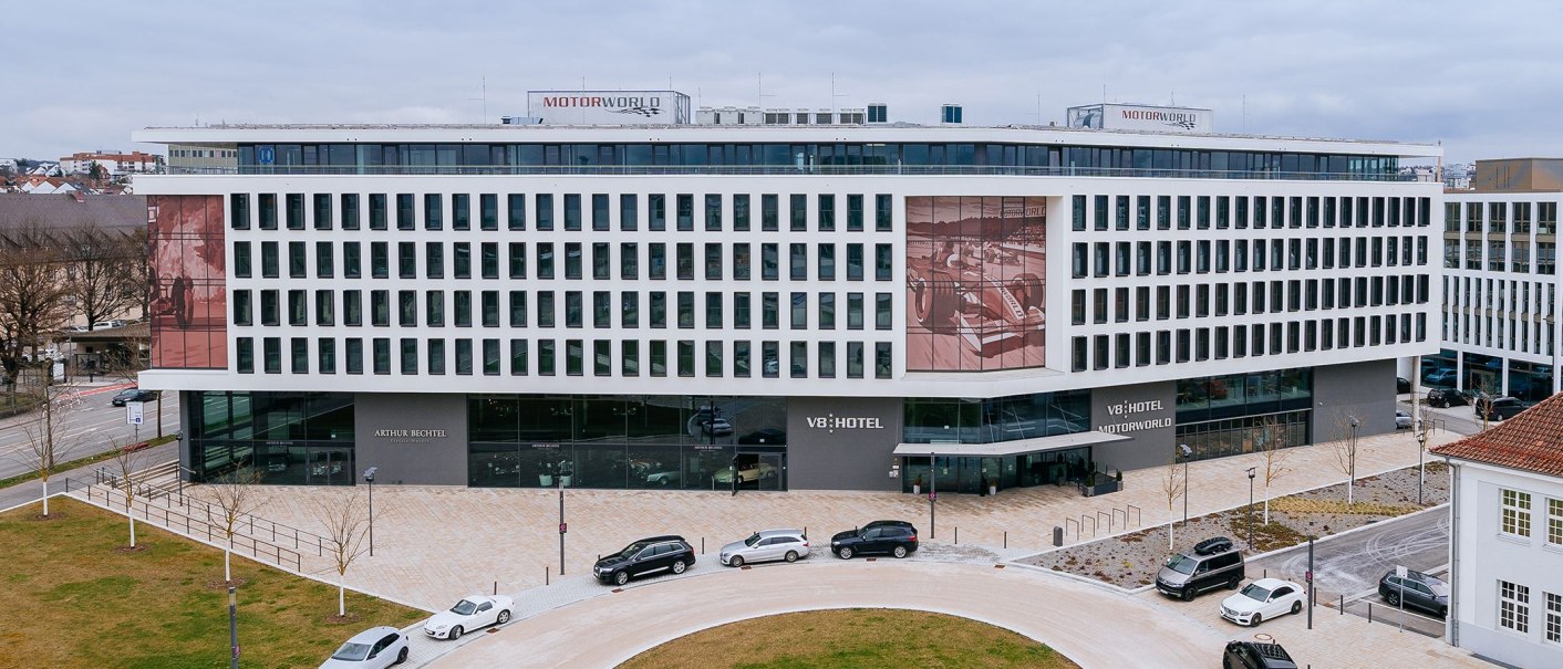 Das V8 HOTEL ist ein modernes Gebäude mit vielen Fenstern. Vor dem Hotel parken mehrere Autos. Auf dem Dach steht "MOTORWORLD"., © V8 HOTEL Das V8 HOTEL ist ein modernes Gebäude mit vielen Fenstern. Vor dem Hotel parken mehrere Autos. Auf dem Dach steht "MOTORWORLD"., © V8 HOTEL