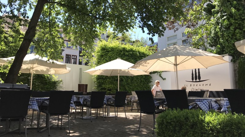 Ein sonniger Garten mit Tischen, St&uuml;hlen und Sonnenschirmen, umgeben von B&auml;umen und Pflanzen. Im Hintergrund ist eine Wand mit der Aufschrift 'Il Boccone' sichtbar., &copy; Restaurant Il Boccone