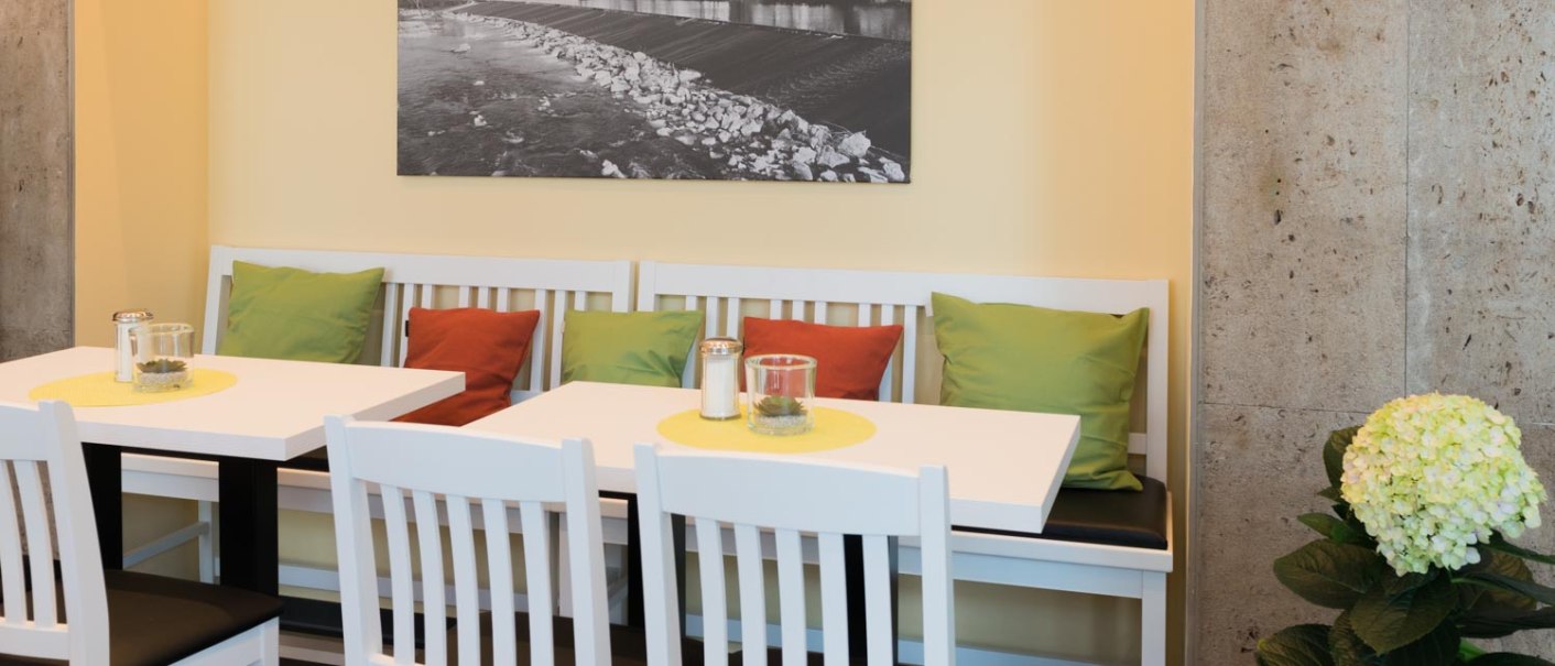 Café mit weißen Tischen und Stühlen, bunten Kissen und einem Schwarz-Weiß-Foto an der Wand. Eine Pflanze steht in der Ecke., © Bäckerei Veit, Dr. Michael Immendörfer