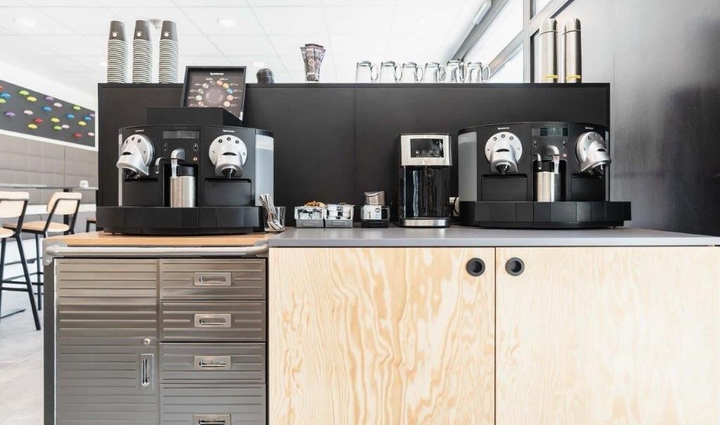 Moderne Kaffestation mit zwei Kaffeemaschinen, Bechern und Zubeh&ouml;r auf einem Holzschrank in einem hellen Raum., &copy; a2 HOTELS am Quadrium