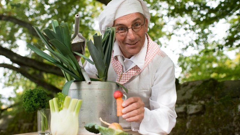 Ein Mann in Kochkleidung mit Brille hält Gemüse in einem Garten. Er hat eine Möhre in der Hand und lächelt freundlich., © Paavo Blofeld Ein Mann in Kochkleidung mit Brille hält Gemüse in einem Garten. Er hat eine Möhre in der Hand und lächelt freundlich., © Paavo Blofeld