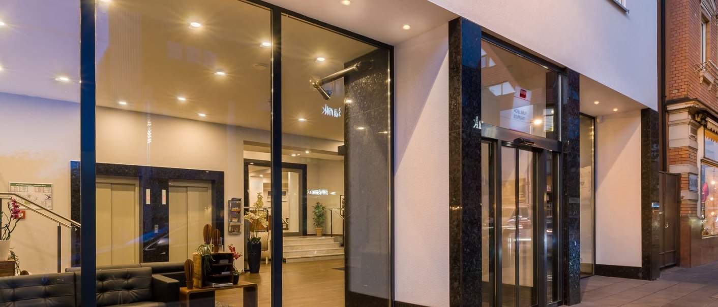 Moderner Eingang mit großer Glasfront, beleuchtetem Innenbereich und eleganter Einrichtung. Außenbereich mit gepflastertem Gehweg., © Novum Hospitality