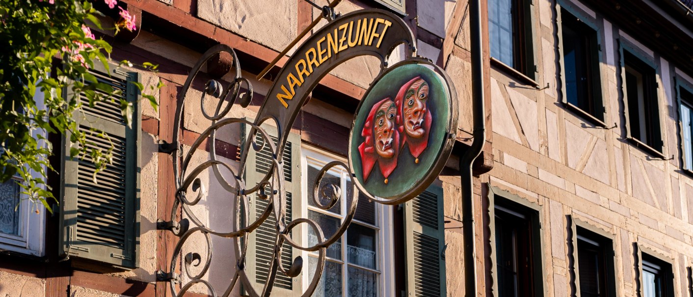 Ein kunstvoll gestaltetes Schild der Narrenzunft hängt an einem Fachwerkhaus in Weil der Stadt. Es zeigt zwei Masken mit roten Kopftüchern., © Olivia + RichiPhotoart Ein kunstvoll gestaltetes Schild der Narrenzunft hängt an einem Fachwerkhaus in Weil der Stadt. Es zeigt zwei Masken mit roten Kopftüchern., © Olivia + RichiPhotoart
