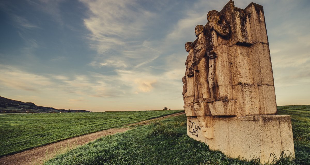 Steinskulptur mit mehreren Figuren auf einem Feldweg. Der Himmel ist leicht bewölkt, die Landschaft weitläufig und grün. Sonnenuntergangsstimmung., © Landratsamt Ostalbkreis Steinskulptur mit mehreren Figuren auf einem Feldweg. Der Himmel ist leicht bewölkt, die Landschaft weitläufig und grün. Sonnenuntergangsstimmung., © Landratsamt Ostalbkreis