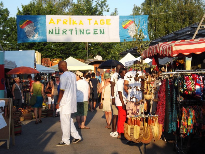 Besucher schlendern über die Afrika Tage Nürtingen, umgeben von bunten Marktständen und einem Banner im Hintergrund., © Stadt Nürtingen Besucher schlendern über die Afrika Tage Nürtingen, umgeben von bunten Marktständen und einem Banner im Hintergrund., © Stadt Nürtingen