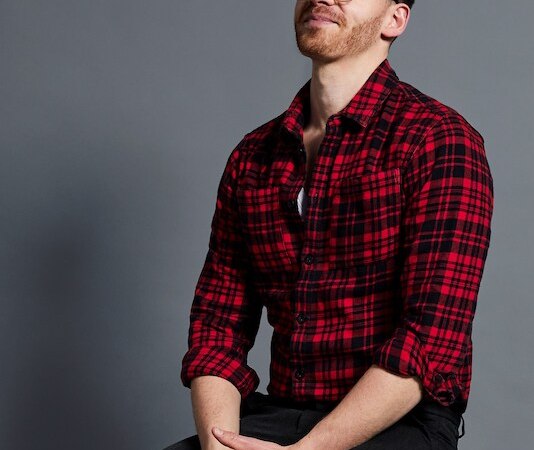 Mann in rotem Karohemd sitzt auf einem Hocker vor grauem Hintergrund, lächelt leicht und schaut nach oben., © Konzertagentur Jochen Dietz