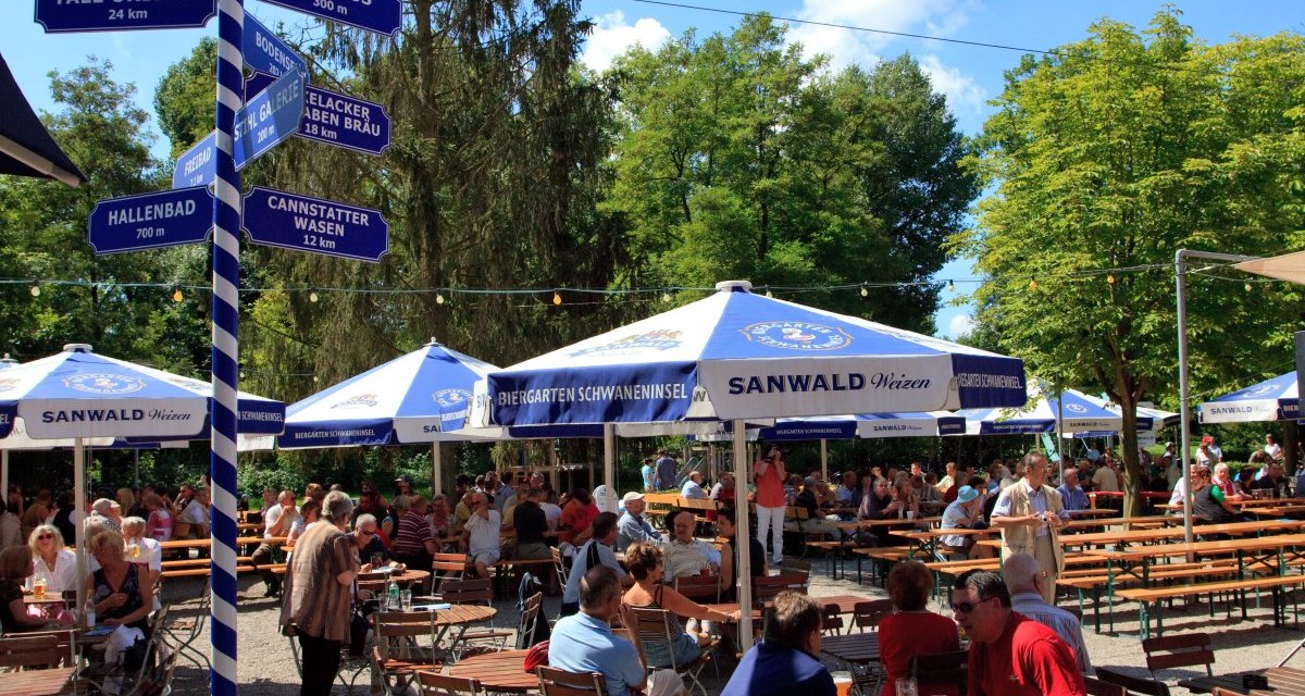 Ein belebter Biergarten mit Holztischen und -stühlen, blauen Sonnenschirmen und einem Wegweiser. Menschen genießen das sonnige Wetter., © Waiblingen - Stuttgart-Marketing GmbH Ein belebter Biergarten mit Holztischen und -stühlen, blauen Sonnenschirmen und einem Wegweiser. Menschen genießen das sonnige Wetter., © Waiblingen - Stuttgart-Marketing GmbH