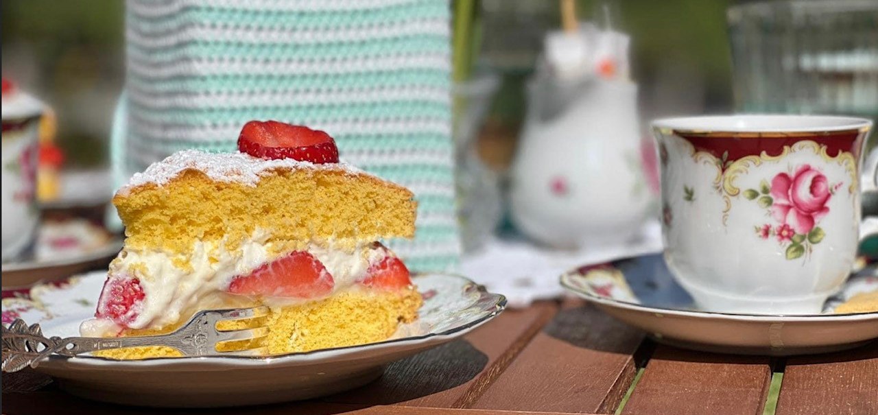 Ein Stück Erdbeerkuchen und eine Tasse Kaffee auf einem Holztisch im Freien. Im Hintergrund eine Tulpe und ein gehäkelter Überzug., © Café Plätscherwiese