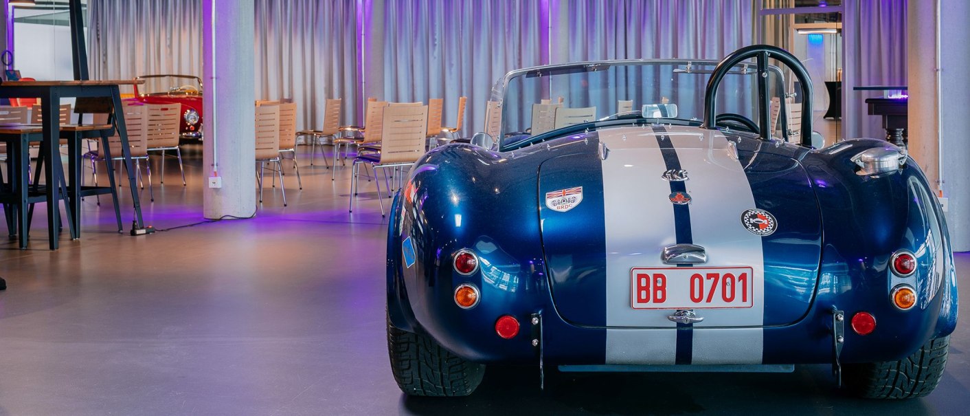 Ein blauer Oldtimer mit weißen Streifen steht in einem modernen Tagungsraum mit Tischen und Stühlen., © V8 HOTEL Ein blauer Oldtimer mit weißen Streifen steht in einem modernen Tagungsraum mit Tischen und Stühlen., © V8 HOTEL