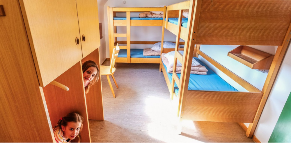 Zwei Etagenbetten in einem hellen Raum, zwei Personen schauen lächelnd aus einem Schrank hervor., © DJH Baden-Württemberg e.V.