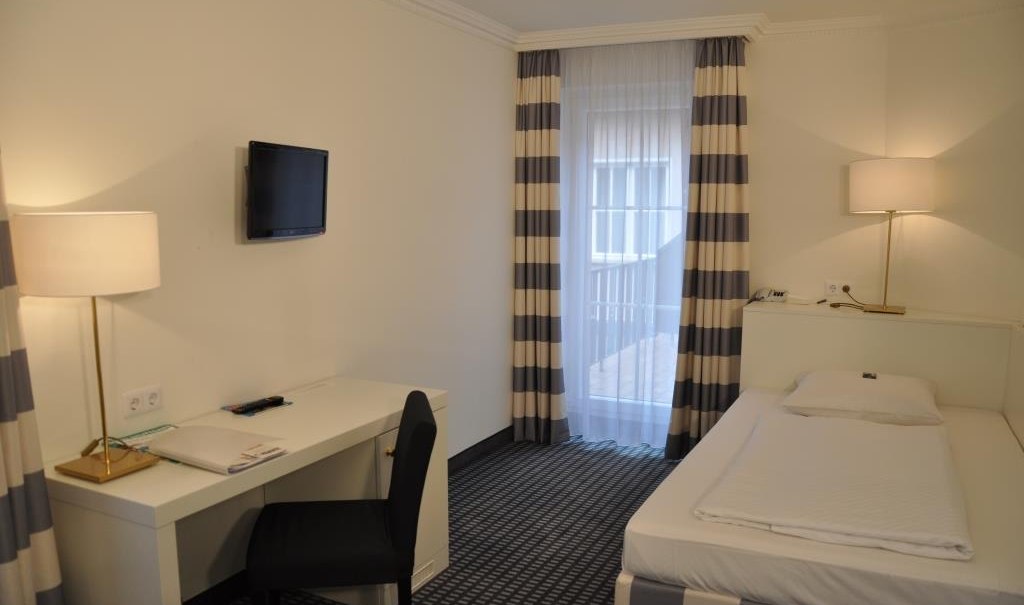 Helles Einzelzimmer mit Bett, Schreibtisch, Stuhl und Wandfernseher. Große Fenster mit gestreiften Vorhängen und zwei Stehlampen., © relexa Waldhotel Schatten Helles Einzelzimmer mit Bett, Schreibtisch, Stuhl und Wandfernseher. Große Fenster mit gestreiften Vorhängen und zwei Stehlampen., © relexa Waldhotel Schatten