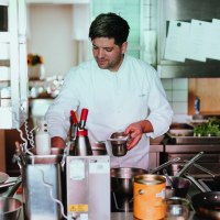 Ein Koch in weißer Uniform arbeitet konzentriert in einer professionellen Küche mit Töpfen und Utensilien., © Daniela Reske