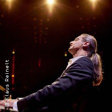 Ein Mann mit langen Haaren spielt Klavier auf einer Bühne, umgeben von warmem Licht., © links im Bild