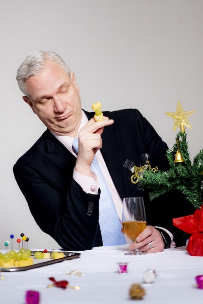 Ein Mann in Anzug hält ein Getränk und eine gelbe Spielfigur. Vor ihm steht ein Mini-Weihnachtsbaum und ein Tablett mit Snacks., © Stadthalle Leonberg Ein Mann in Anzug hält ein Getränk und eine gelbe Spielfigur. Vor ihm steht ein Mini-Weihnachtsbaum und ein Tablett mit Snacks., © Stadthalle Leonberg