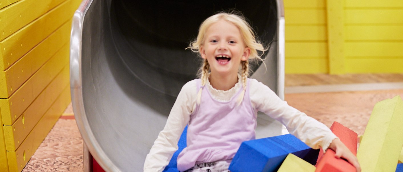 Ein Kind rutscht lachend aus einer orangefarbenen Röhre auf einer Kinderbaustelle. Umgeben von bunten Schaumstoffblöcken in Gelb, Blau und Rot., © Julia Ochs Ein Kind rutscht lachend aus einer orangefarbenen Röhre auf einer Kinderbaustelle. Umgeben von bunten Schaumstoffblöcken in Gelb, Blau und Rot., © Julia Ochs