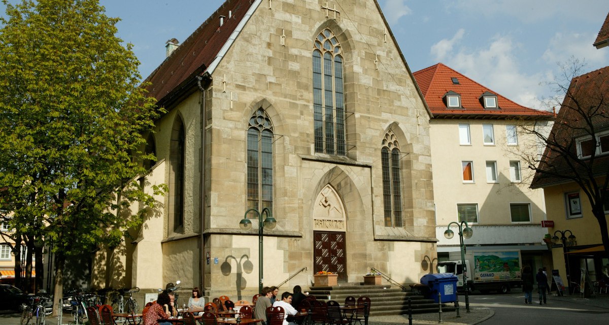 Die Nikolaikirche mit gotischen Fenstern, umgeben von Bäumen und Sitzgelegenheiten. Menschen sitzen draußen bei sonnigem Wetter., © Sphäre Verlag