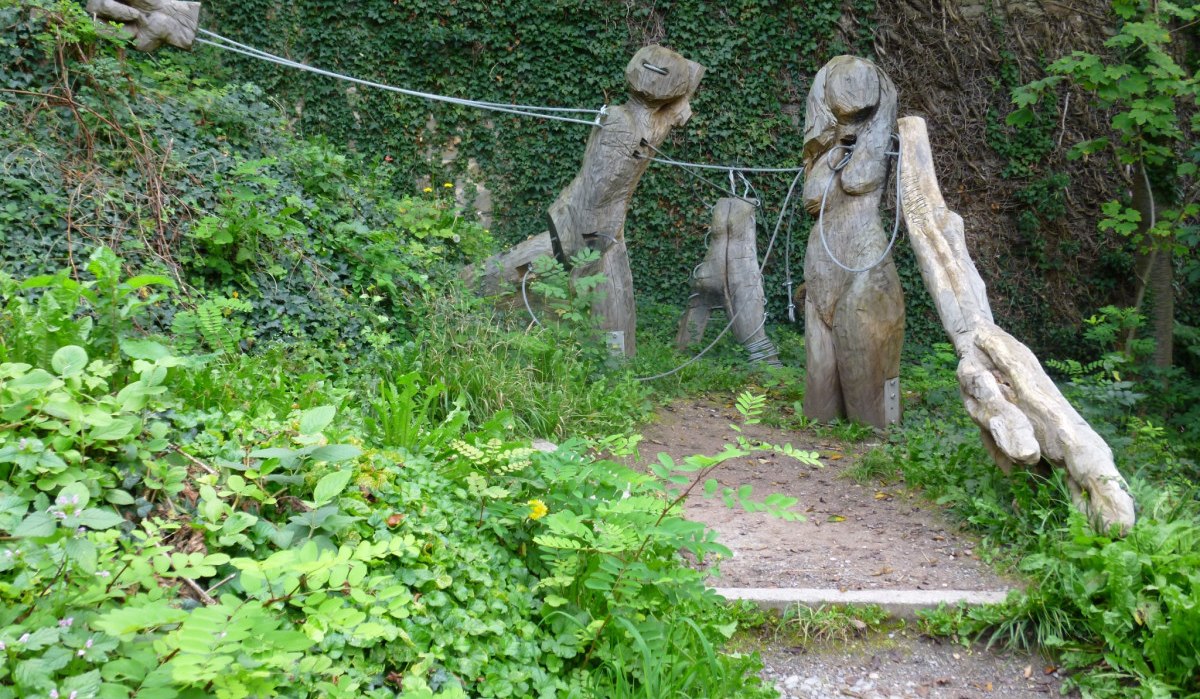 Holzskulpturen auf einem Pfad, umgeben von &uuml;ppigem Gr&uuml;n und Kletterpflanzen. Die Figuren sind durch Seile verbunden., &copy; Natur.Nah. Sch&ouml;nbuch & Heckeng&auml;u