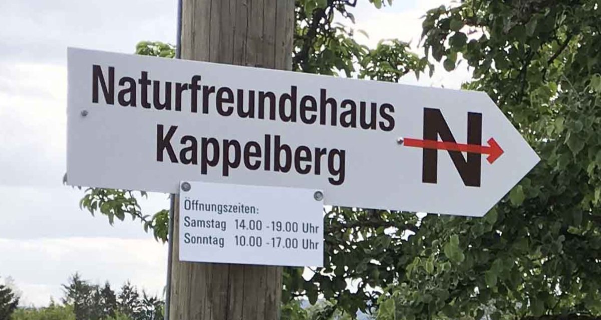 Wegweiser zum Naturfreundehaus Kappelberg mit Öffnungszeiten für Samstag und Sonntag, montiert an einem Holzpfosten vor Bäumen. Wegweiser zum Naturfreundehaus Kappelberg mit Öffnungszeiten für Samstag und Sonntag, montiert an einem Holzpfosten vor Bäumen.