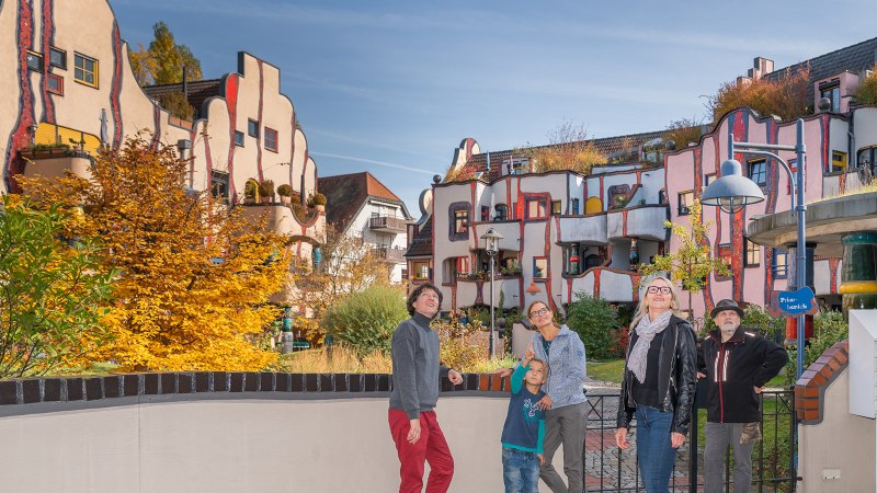 Menschen stehen vor dem farbenfrohen Hundertwasser-Wohnhaus in Plochingen. Die Fassade ist wellenförmig und bunt gestaltet, umgeben von Herbstbäumen., © Bildergalerie Attilla Menschen stehen vor dem farbenfrohen Hundertwasser-Wohnhaus in Plochingen. Die Fassade ist wellenförmig und bunt gestaltet, umgeben von Herbstbäumen., © Bildergalerie Attilla