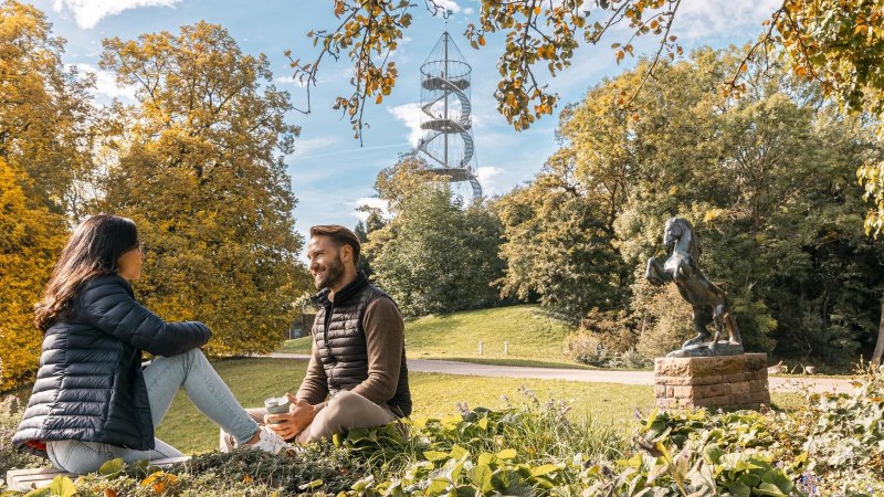 Zwei Personen sitzen im H&ouml;henpark Killesberg, umgeben von Herbstb&auml;umen. Im Hintergrund ist der Killesbergturm zu sehen, neben einer Pferdestatue., &copy; Stuttgart-Marketing GmbH, Sarah Schmid