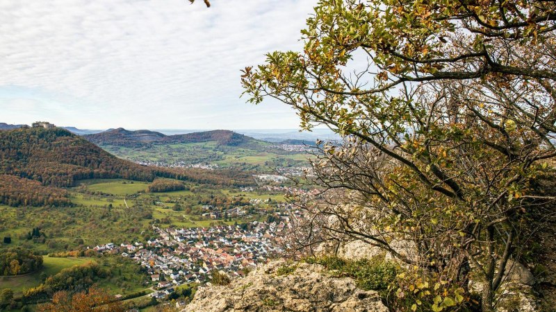 Panoramablick vom Beurener Fels über ein grünes Tal mit einem Dorf und bewaldeten Hügeln im Hintergrund. Ein Wanderer steht am Rand des Felsens., © Stuttgart-Marketing GmbH, Sarah Schmid