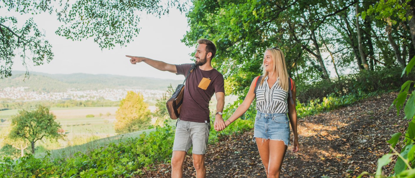 Ein Paar wandert im Remstal, hält sich an den Händen. Der Mann zeigt in die Ferne. Sie sind von grünen Bäumen umgeben, im Hintergrund eine weite Landschaft., © Bebop Media