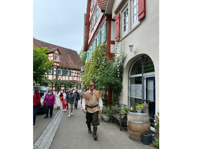 Ein Mann in historischer Kleidung geht durch eine malerische Straße in Schorndorf, flankiert von Fachwerkhäusern und einer Gruppe von Besuchern., © Eigenbetrieb Tourismus und Citymanagement Schorndorf Ein Mann in historischer Kleidung geht durch eine malerische Straße in Schorndorf, flankiert von Fachwerkhäusern und einer Gruppe von Besuchern., © Eigenbetrieb Tourismus und Citymanagement Schorndorf