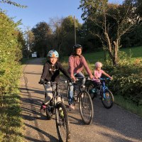 Drei Personen auf Fahrrädern fahren bei sonnigem Wetter auf einem Radweg durch eine grüne Landschaft., © T&M