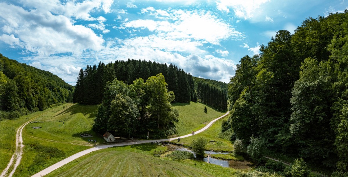 Vogelperspektive auf eine gr&uuml;ne Landschaft mit W&auml;ldern, Wiesen und einem kleinen Bach. Ein Weg schl&auml;ngelt sich durch die Szenerie unter einem blauen Himmel., &copy; Landkreis G&ouml;ppingen