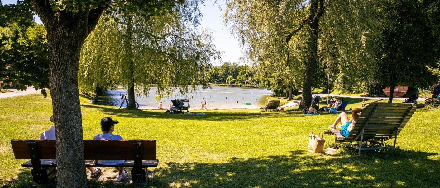 Menschen genießen einen sonnigen Tag am Waldsee in Fornsbach. Einige sitzen auf Bänken, andere liegen auf der Wiese. Der See und Bäume bieten eine idyllische Kulisse., © Stuttgart-Marketing GmbH, Sarah Schmid Menschen genießen einen sonnigen Tag am Waldsee in Fornsbach. Einige sitzen auf Bänken, andere liegen auf der Wiese. Der See und Bäume bieten eine idyllische Kulisse., © Stuttgart-Marketing GmbH, Sarah Schmid