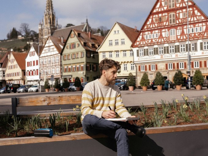 Ein Mann skizziert auf einer Bank in Esslingen, umgeben von historischen Fachwerkhäusern und einer Kirche im Hintergrund., © Esslinger Stadtmarketing &amp; Tourismus GmbH