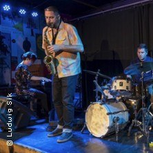 Eine Jazzband spielt auf einer B&uuml;hne. Der Saxophonist steht im Vordergrund, w&auml;hrend der Pianist und der Schlagzeuger im Hintergrund zu sehen sind., &copy; links im Bild