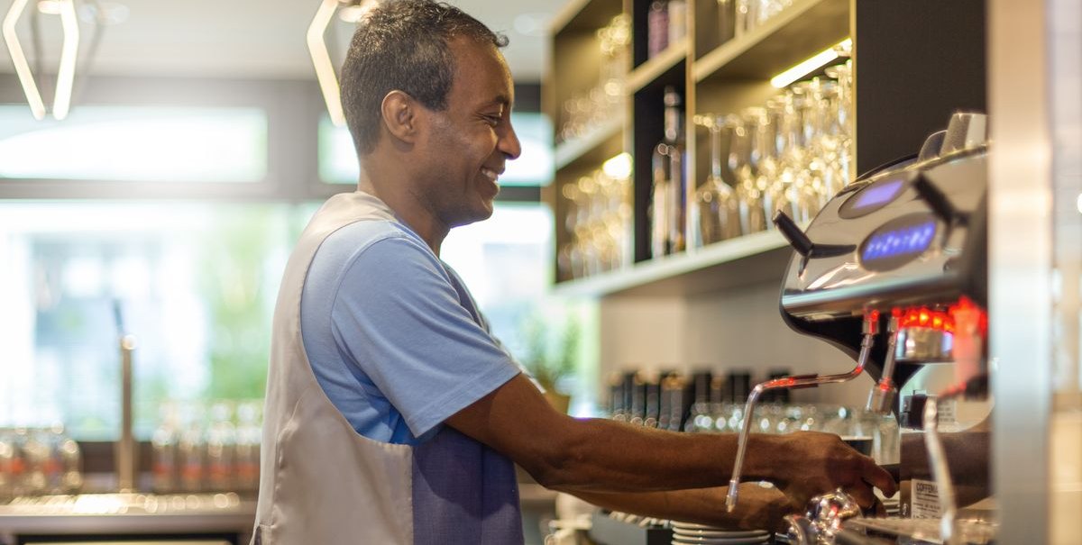 Ein Mann arbeitet lächelnd an einer Espressomaschine in einer modernen Bar mit Gläsern im Hintergrund., © Novum Hospitality Ein Mann arbeitet lächelnd an einer Espressomaschine in einer modernen Bar mit Gläsern im Hintergrund., © Novum Hospitality